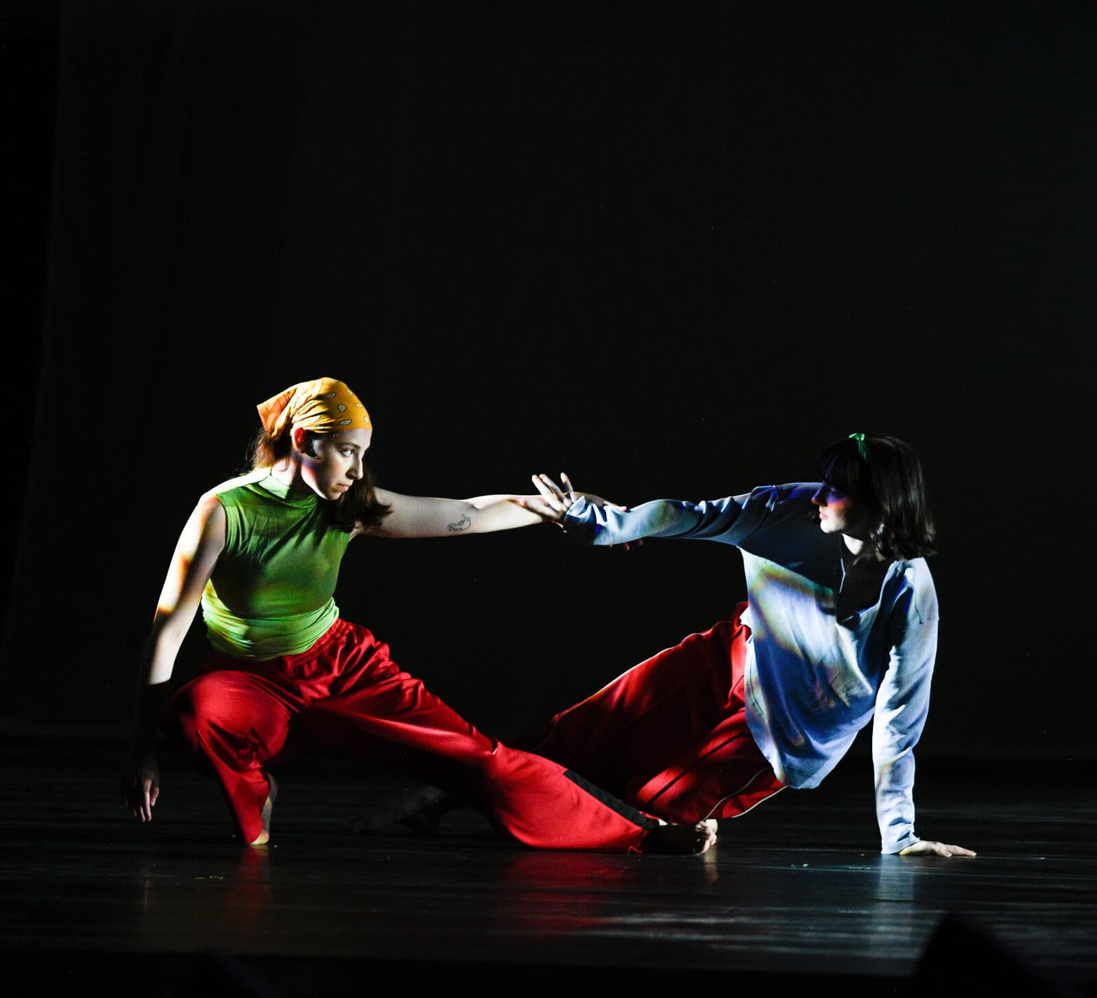 Two dancers in bright costumes reach toward each other across a dark stage, fingertips almost meeting.