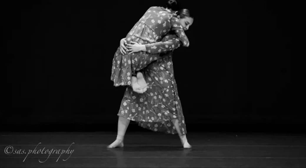 Black and white photograph of two dancers in matching floral dresses on a dark stage, one folded over the other in an embrace.