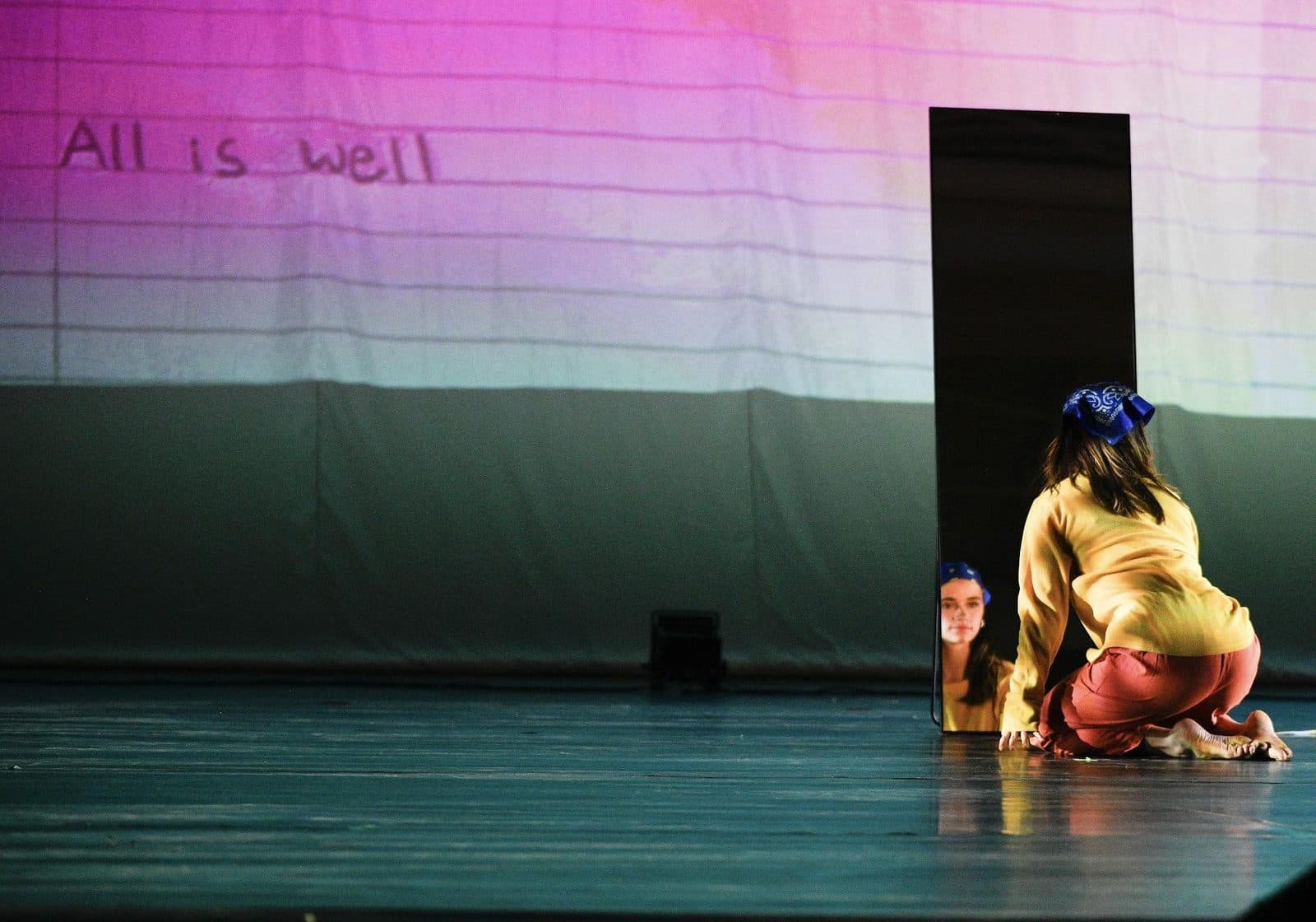 A dancer kneels before a tall standing mirror, her reflection meeting her, as the words “All is well” are projected across the backdrop behind her.