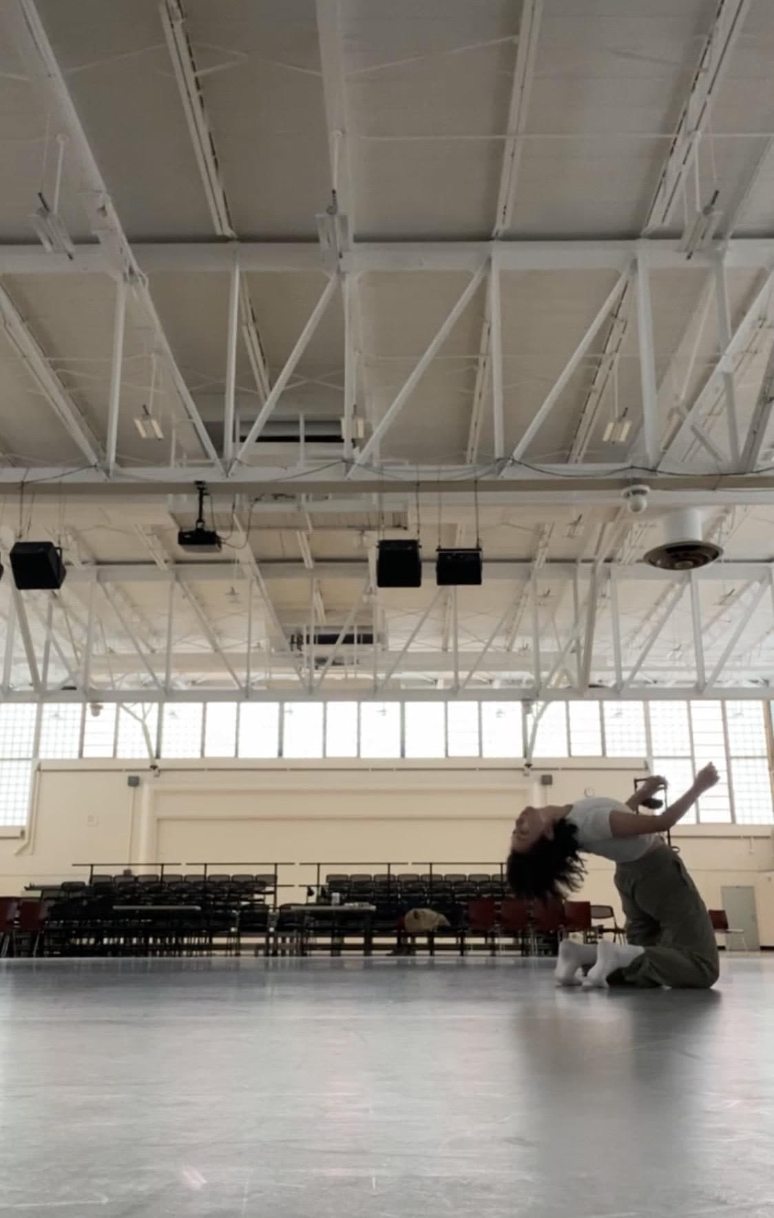 A dancer on her knees in a bright, empty studio, arching backward with one arm thrown overhead.