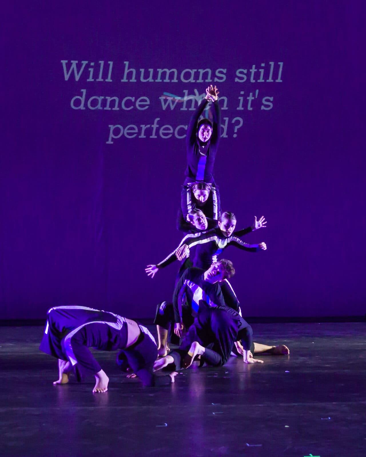 Dancers in stick-figure costumes stacked into a human pyramid under violet light, beneath the projected question “Will humans still dance when it’s perfect?”