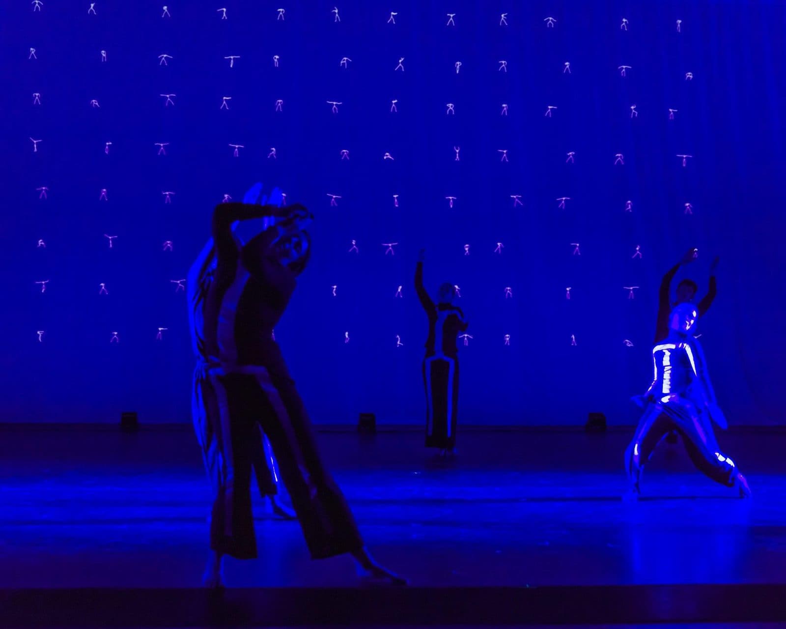 Three dancers on a stage washed in deep blue, before a backdrop projected with rows of small stick figures.