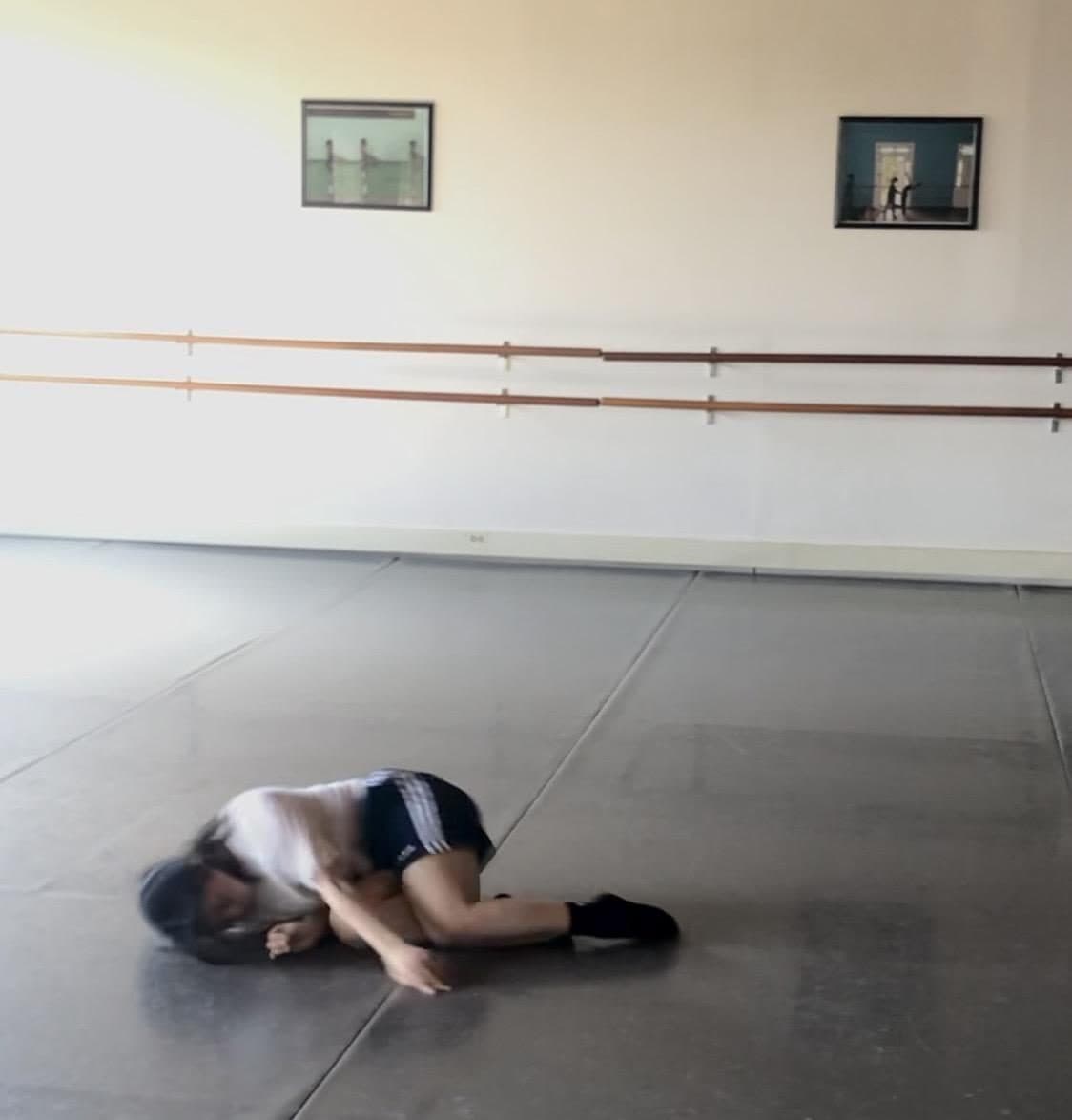 A dancer curled on her side on the floor of a ballet studio, barres and framed prints on the wall behind her.